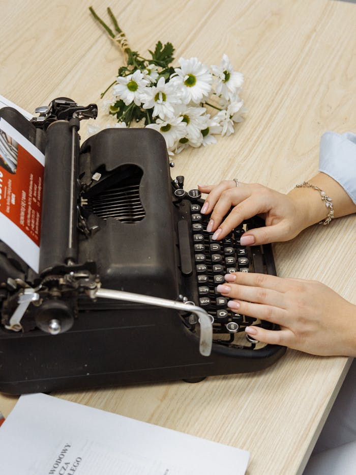 A vintage typewriter with a woman's hands typing, surrounded by flowers on a wooden table.