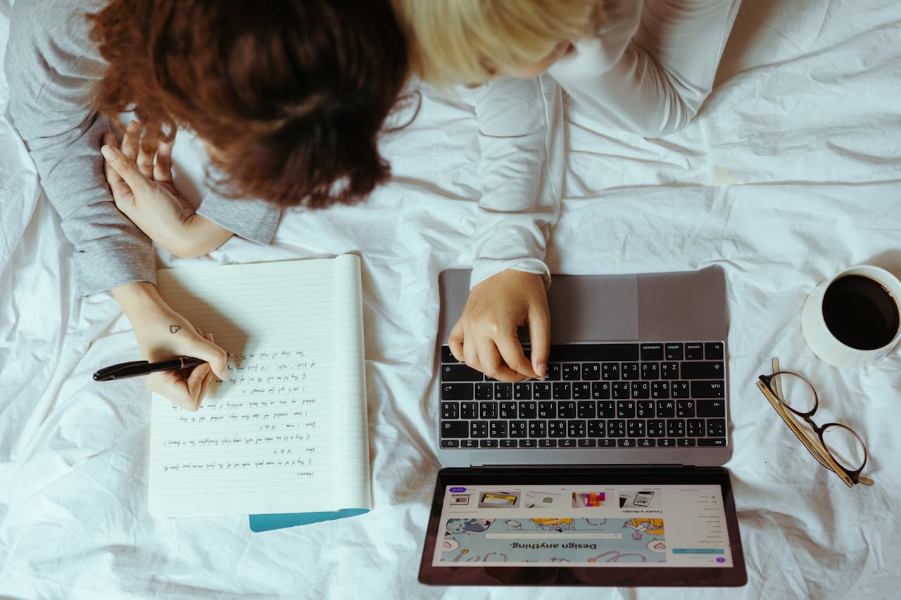 Top view of women lying on bed with laptop and notebook while writing and surfing internet together during work with project