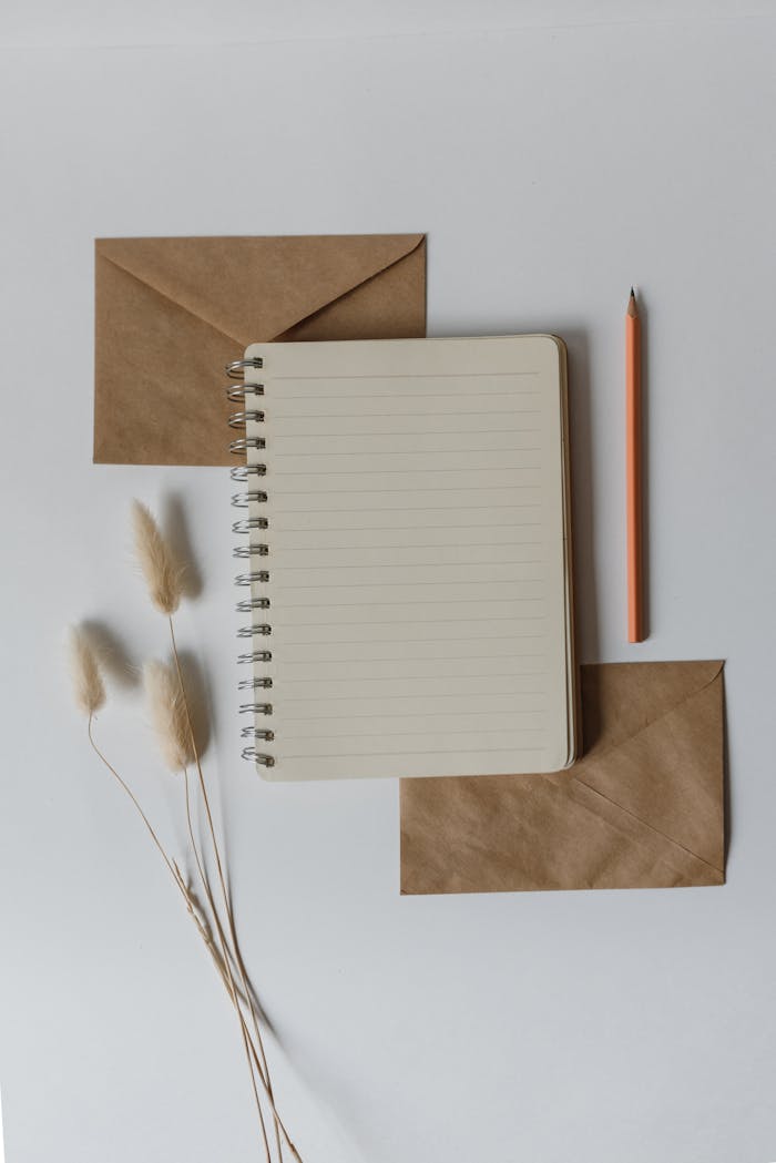 Top view of a spiral notebook with envelopes and a pencil on a white surface.